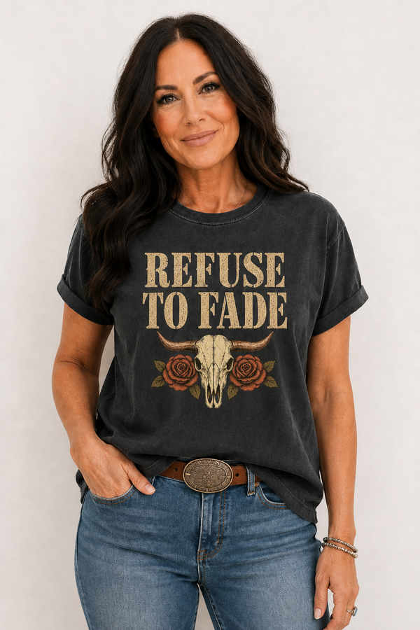 A woman wearing a dark charcoal gray graphic t-shirt with the text 'REFUSE TO FADE' in beige, decorated with a longhorn skull and roses. She is also wearing blue jeans and a western belt, posing against a light gray background.